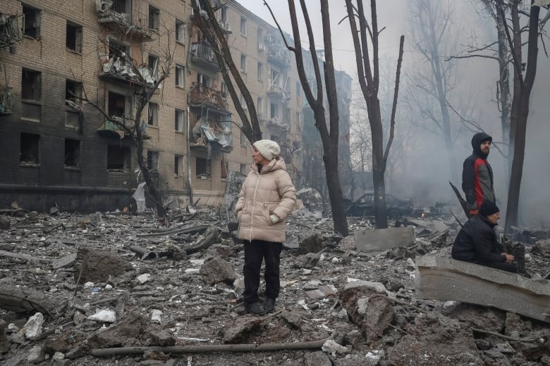 Two men and a woman in winter gear mill about in a pile of rubble next to a charred building with blown-out balconies. Behind them, smoke lingers in the air. 