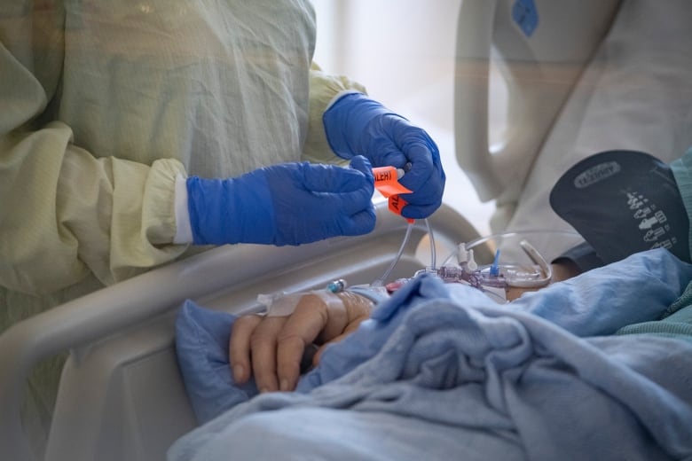 A close-up of a patient's hand as they lay in a medical bed. A health-care worker wearing a gown and gloves attends to them.