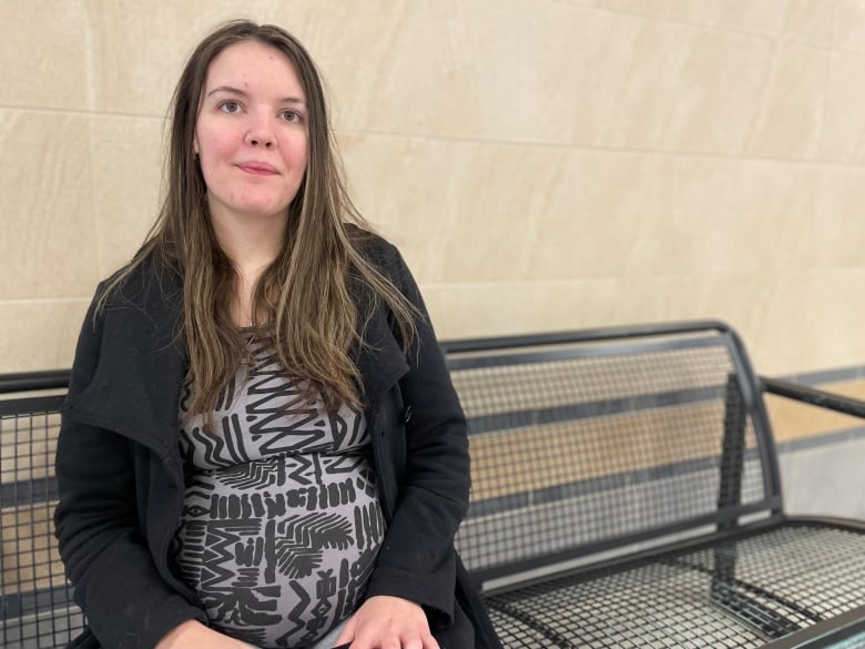 A woman with long hair and a long black coat sits on a bench in a mall.