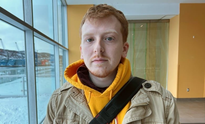 A man in a yellow sweater standing in a lobby near a window. 