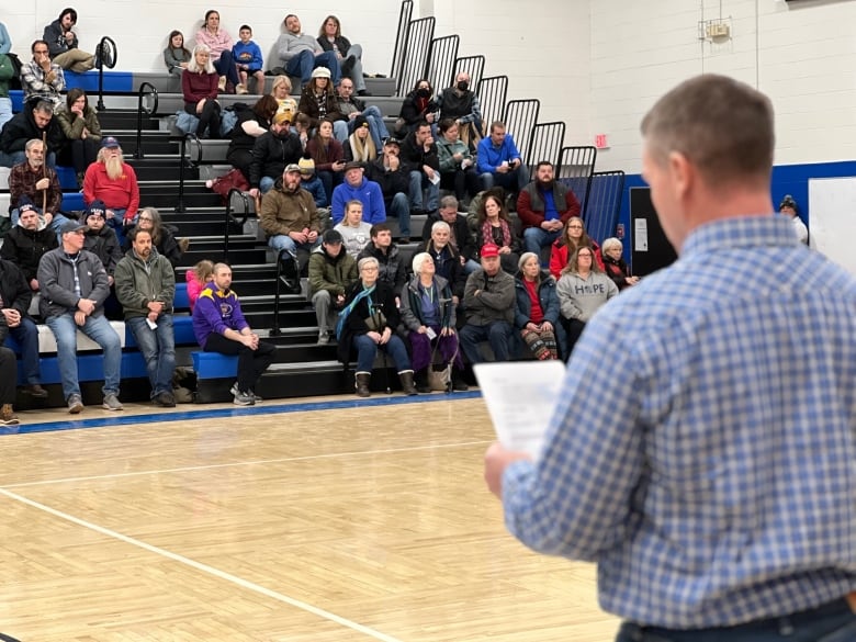 View from behind of man reading from a sheet, speaking to dozens of people seated in the stands of a high-school gym.