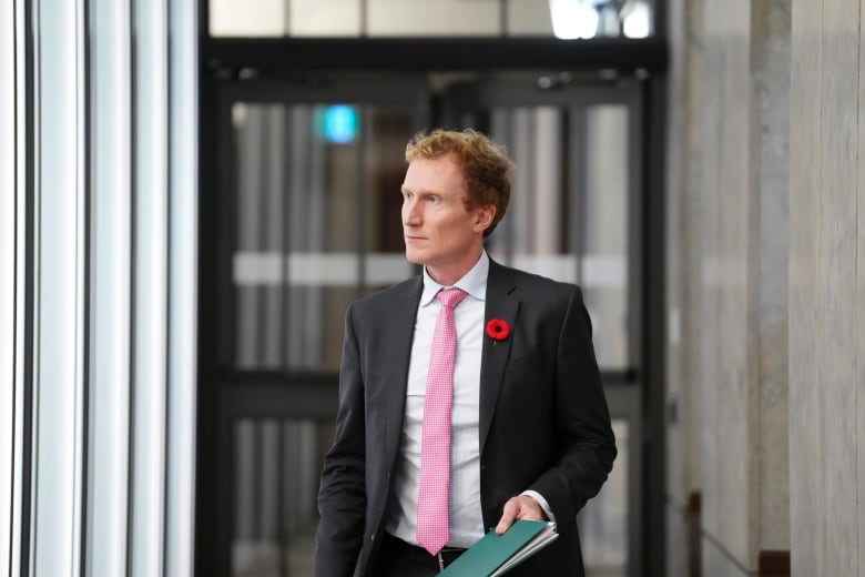 A man in a black suit, white shirt and pink tie walks through the halls of Parliament Hill carrying a green binder.