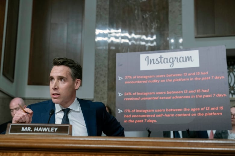 A person gestures while speaking in front of a sign.