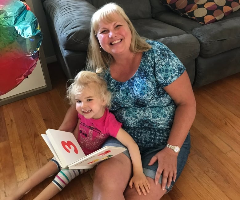 A smiling woman sits on the floor with a smiling girl holding a picture book.