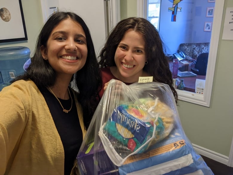 Two smiling women pose for a selfie while holding up a plastic bag filled with menstruation products.  