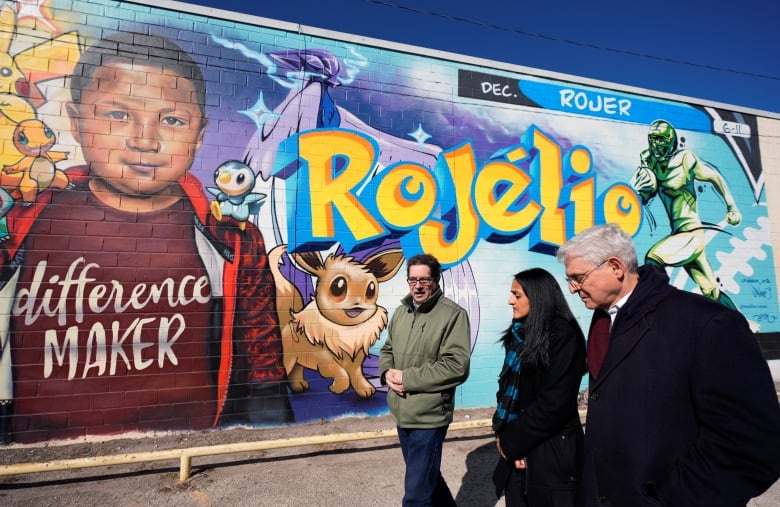 Three people walk past a colourful mural of a boy with the name 'Rojélo' in bright yellow block letters. The boy wears a shirt that says 'Difference maker,' and he's surrounded by Pokemon. On the other side of the mural is a football player, clutching a ball and running.