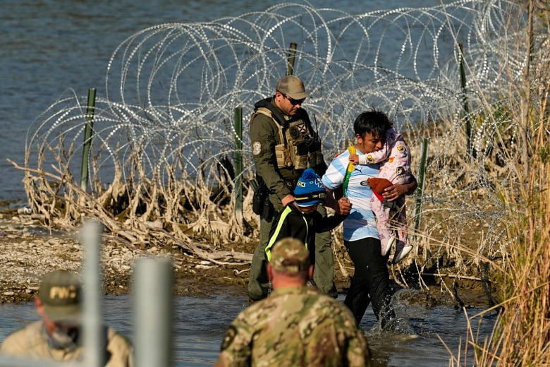 A man carrying a baby wades in shallow water, near barbed wire, with another man in a green uniform following behind.