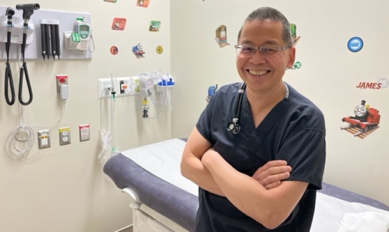 A smiling pediatric doctor in blue scrubs is seen in an unoccupied patient's room adorned with colourful stickers on the walls.