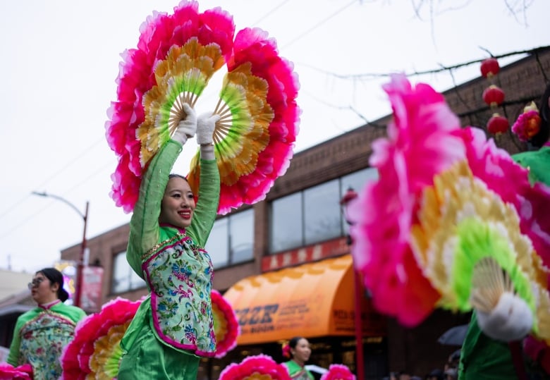 A woman in a pink and green traditional Chinese outfit has her arms stretched up with two pink, yellow and green fans spread open. 