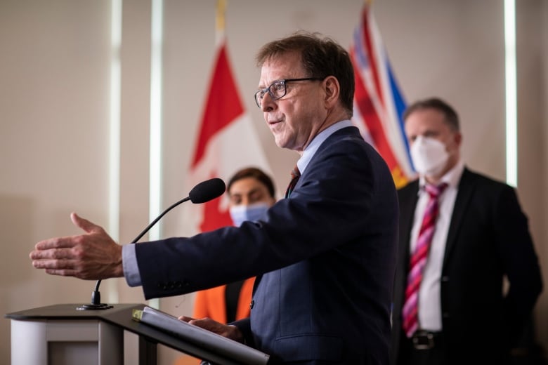 A white man gestures as he speaks at a podium, with two people behind him wearing facemasks.