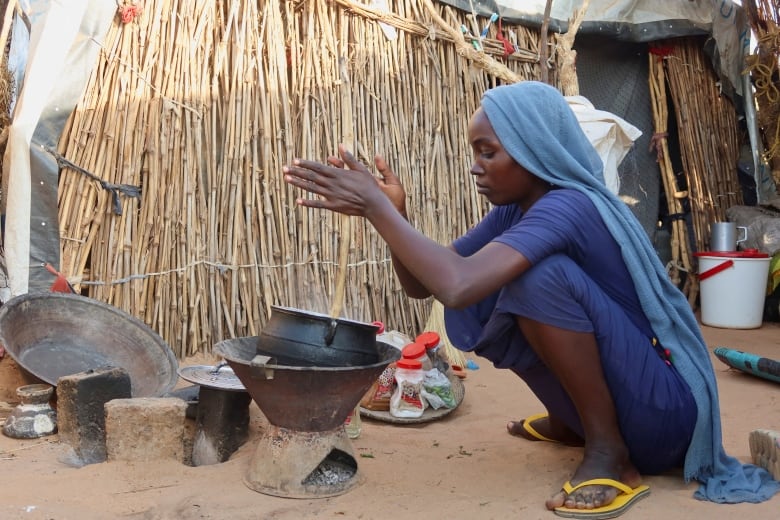 A woman in a long blue dress and headscarf squats in front of a small cooking pot propped in the sand in front of a makeshift bamboo shelter, rolling a stick between her hands. 