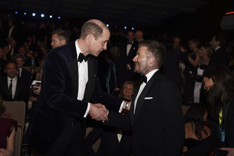 Two people in tuxedos shake hands as people sit in an audience behind then.