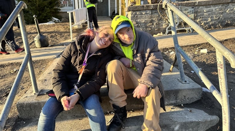 A woman wearing jeans, a black jacket and a purple lanyard sits on a concrete staircase next to a man wearing a neon green hoodie and plaid pants. 