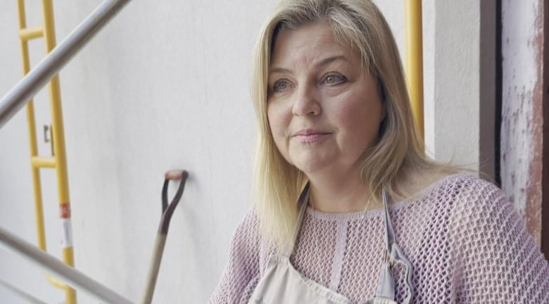 A woman with blonde hair stands outside a restaurant. She's wearing a pink knit sweater and an apron.