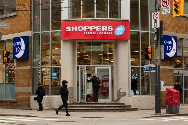 A Shoppers Drug Mart location is pictured at the intersection of on King & Peter streets in downtown Toronto. People wearing winter clothing are walking on the sidewalk in front of the store.