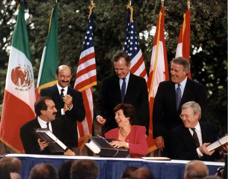 Six people in business suits sit or stand around a table. The American, Mexican and Canadian flags hang in the background.