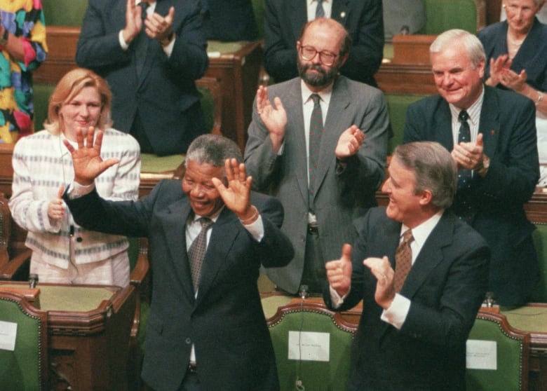 A man in a suit stands and waves with both hands in the air as others around applaud him.