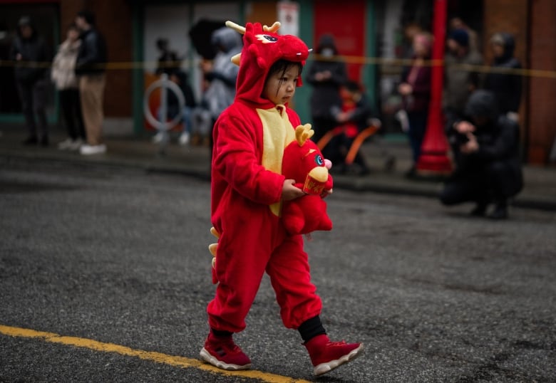 A kid is walking down the street wearing a red dragon onesie and holding a dragon stuffy in her arms.