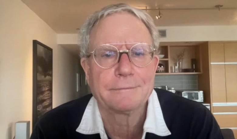 Dr. James Talbot is wearing glasses, a black sweater and white collard shirt as he looks directly into the camera. A kitchen can be seen behind him