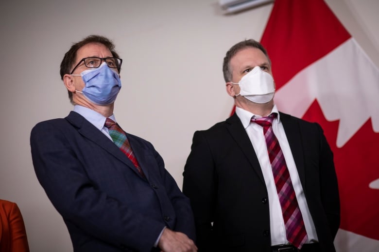 Two white men wearing facemasks stand in front of a Canada flag.