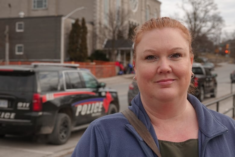 A woman with red hair in a tight ponytail stares. Behind her is a police SUV and a large, stone church buidling.