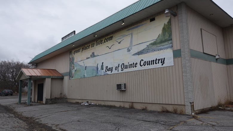 A large, beige building with a massive mural on one wall announcing "Bay of Quinte Country" is a "great place to live." The building is rundown.