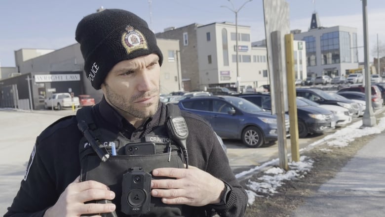 A man weairng a police uniform looks sternly to the right of the frame. He has a toque and his police vest is covered with various tools.