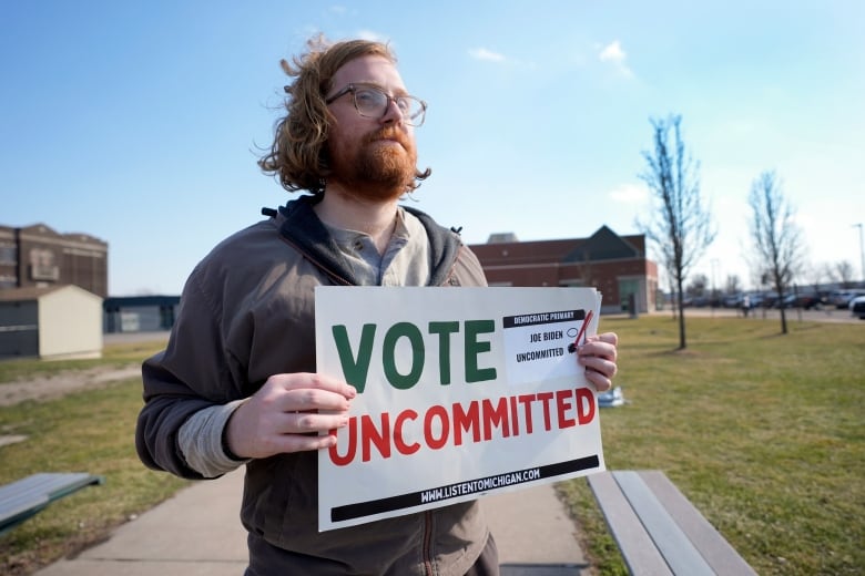 A man stands outside holding a white paper sign that reads "vote uncommitted." 