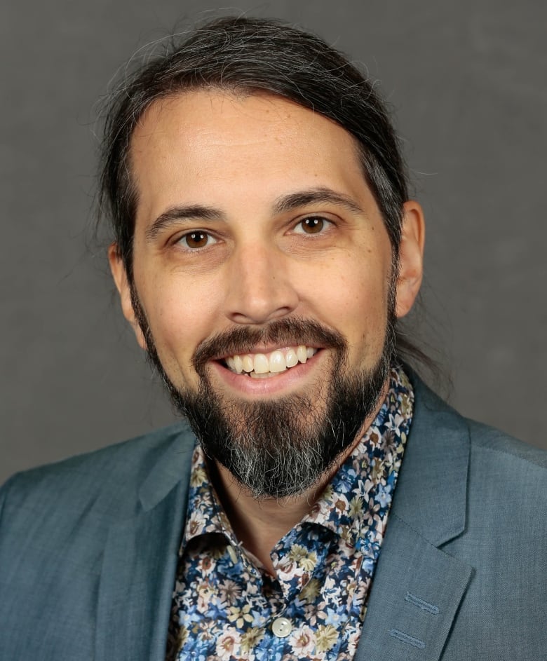 Raiche has a beard and is wearing a blue suit and flowered shirt and is smiling in this headshot