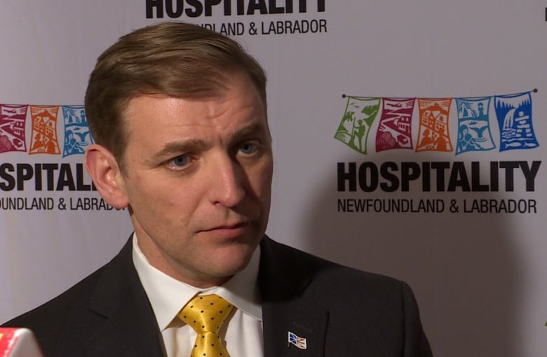 A man wearing a suit and a yellow tie stands in front of a white screen with logos reading, "hospitality newfoundland & labrador."