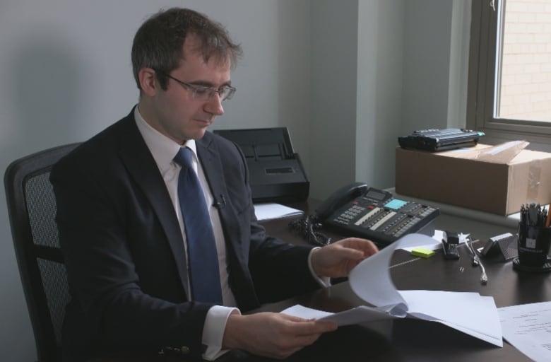 A brown-haired man wearing glasses and a navy suit sits at a desk and looks over papers.