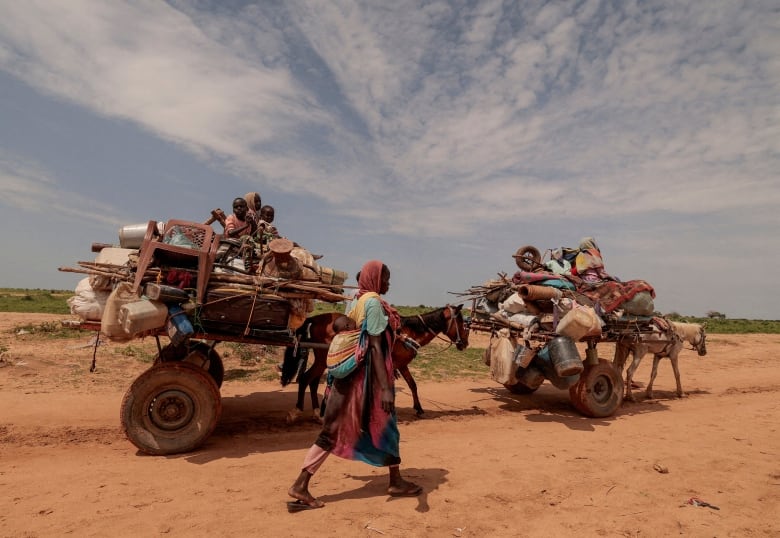 A woman with a baby strapped onto her back walks on a dirt road alongside two rickety carriages, pulled by small horses, carrying families sitting atop piles of belongings tied together with rope. 