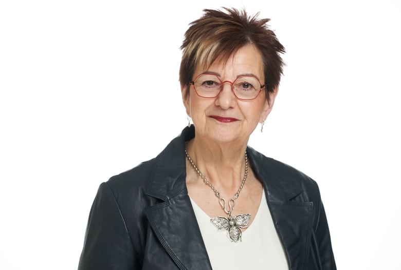 A woman with short, spikey hair and glasses poses for a portrait wearing a black leather blazer and a necklace with a large pendant in the shape of a bee.