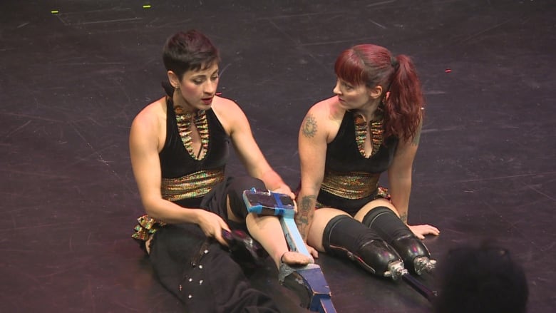 Two people sit on a stage. One has two prosthetic legs; the other has one foot poised on a stilt.