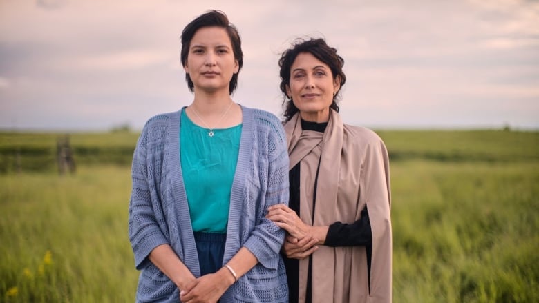 Two women pose in front of a grass field. The woman on the left is younger than the other. The older woman gently rests her hand on the other's arm.