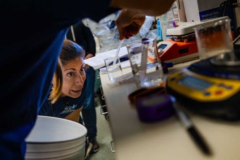 A person is pictured from behind using a plastic syringe to administer drops into a beaker full of clear liquid. Another woman bends down and peers closely at the beaker, her eyebrows raised, her eyes wide open and her mouth agape.  