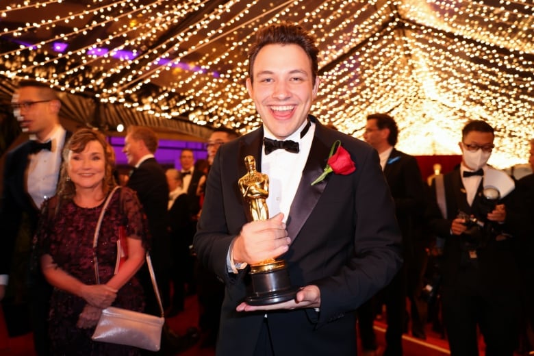 A smiling man wearing a tuxedo with a rose pinned to his lapel holds an Oscar statuette in a crowded room under a canopy of lights.