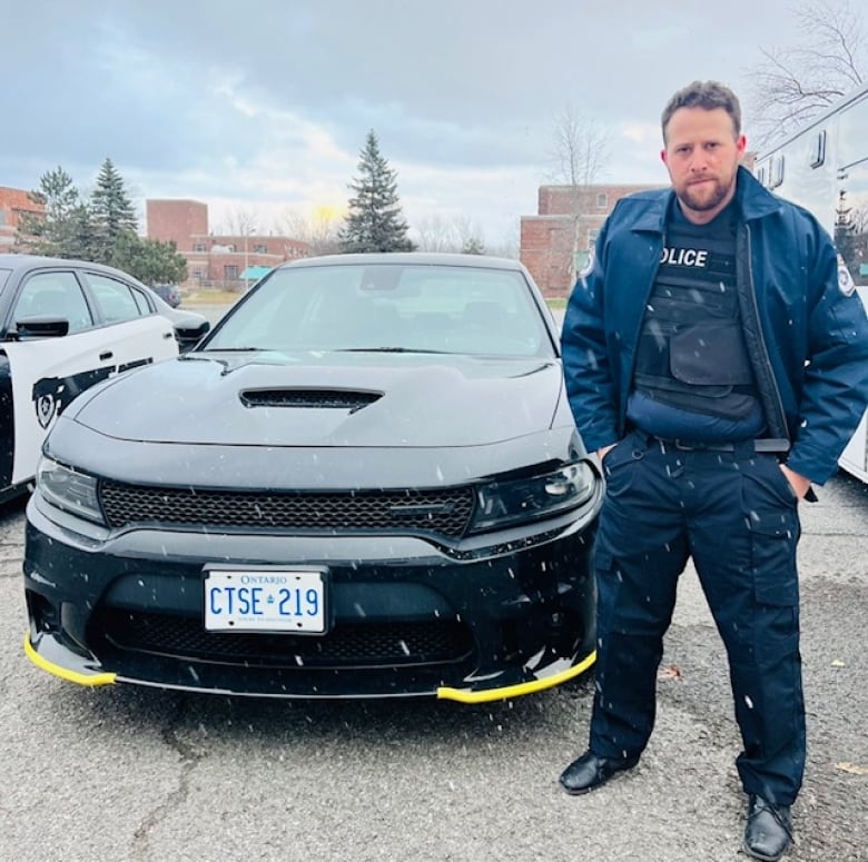 A man wearing a police uniform stands next to a black police car with an Ontario licence plate. 