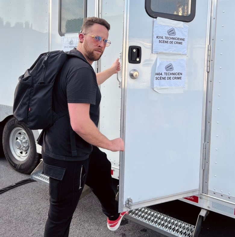 A man wearing a black t-shirt and pants, clear sunglasses, a backpack and red shoes enters a trailer on a film set.