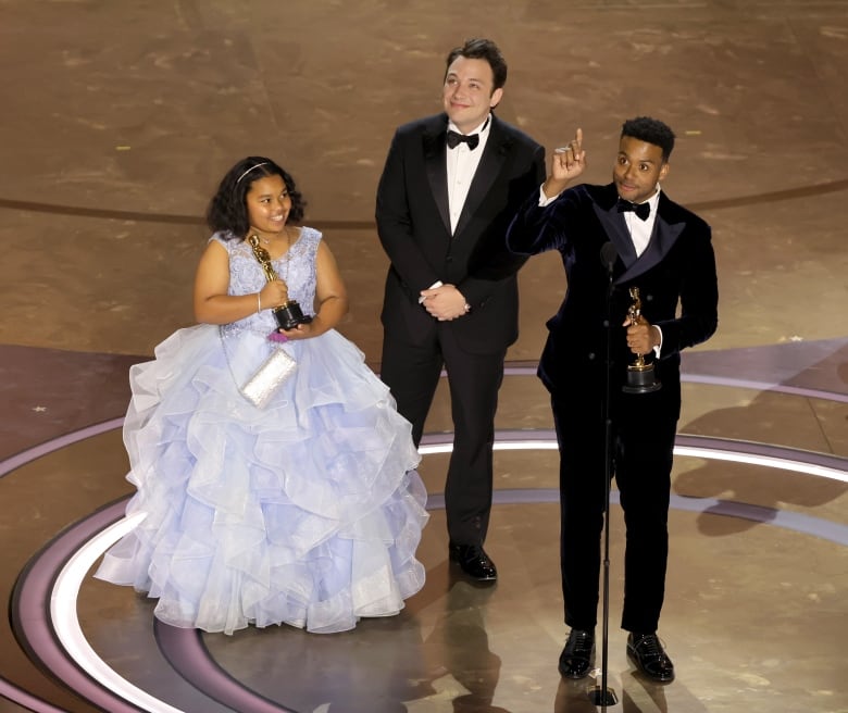 Two men in black suits with bowties stand on a stage next to a young girl in a frilly lavender dress. One man is holding and Oscar and pointing up.