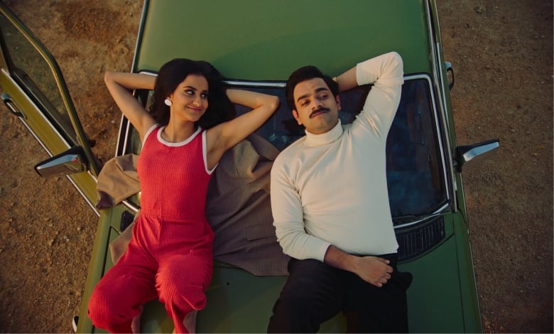 A woman looks at a man while they lie on the hood of a car.