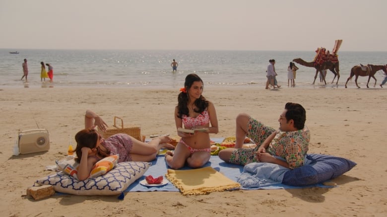 Three people relax on the beach. In the middle, a young woman wearing a white bikini with pink polka dots looks up from a book.