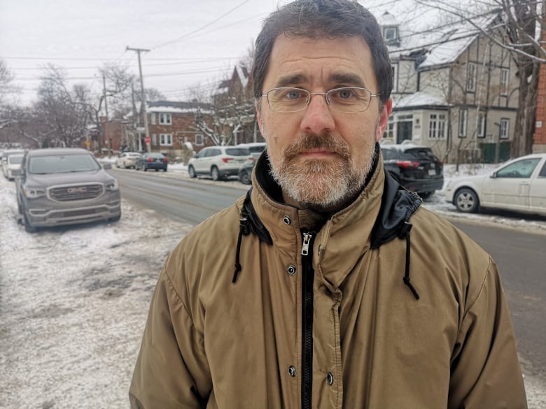 A man with black hair, a grey beard and glasses standing on a snowy, city street.