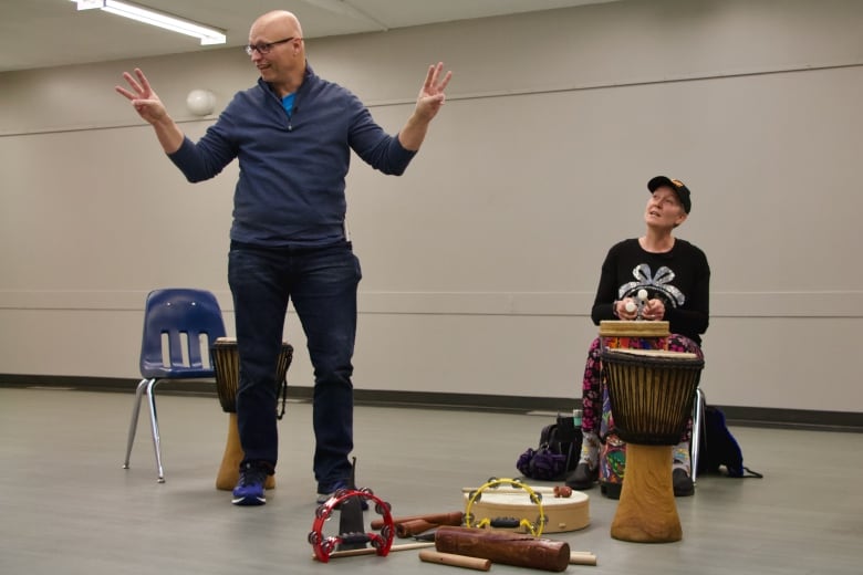 A man stands in a plain room holding up three fingers on each hand. At his feet is an assortment of percussive instruments. A woman sitting at a tall drum looks up at him.