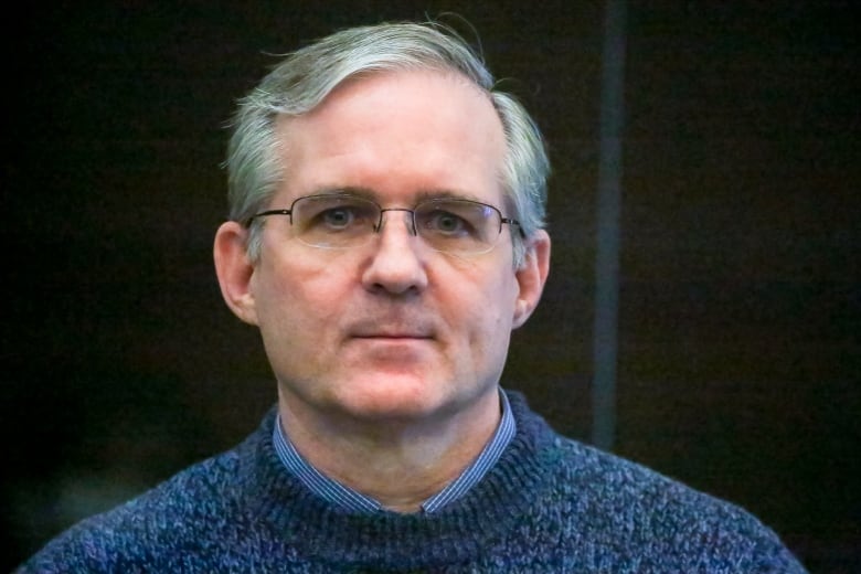 A bespectacled, clean-shaven man wearing a sweater and a collared shirt underneath is shown in a closeup photograph.