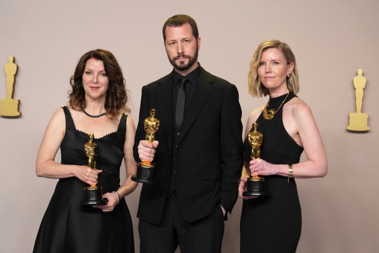 Three people pose on the red carpet holding gold statuettes.