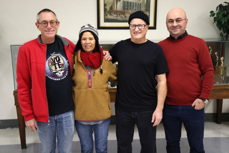 Three men and one woman stand side-by-side, smiling, with their arms around each other's shoulders. 