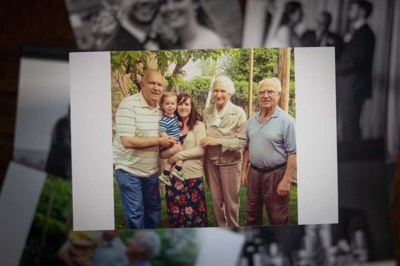 Four generations of a family stand in a garden. A mother hold her little boy next to a grandfather and great grandparents.