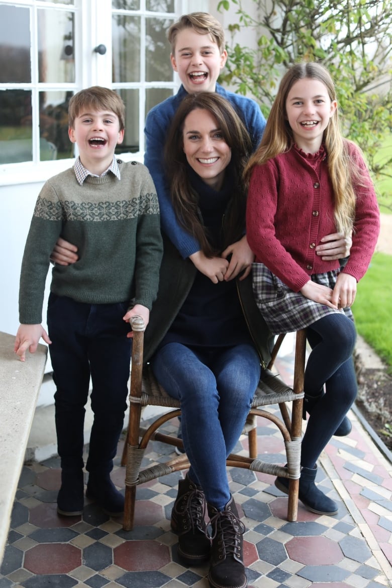 Catherine sits in a chair with two children standing on either side and one behind them. All are smiling. At a first glance and without zooming in nothing appears wrong with the photo.