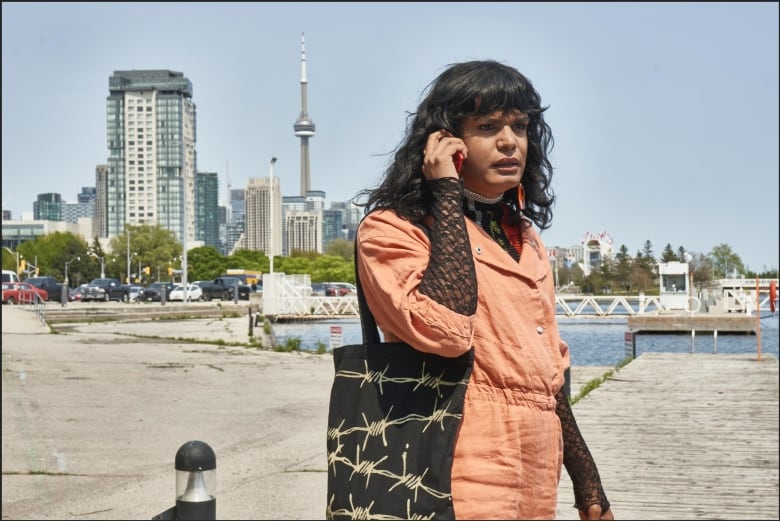 A person standing on a dock holds a cellphone to their ear. Behind them can be seen the CN Tower. 